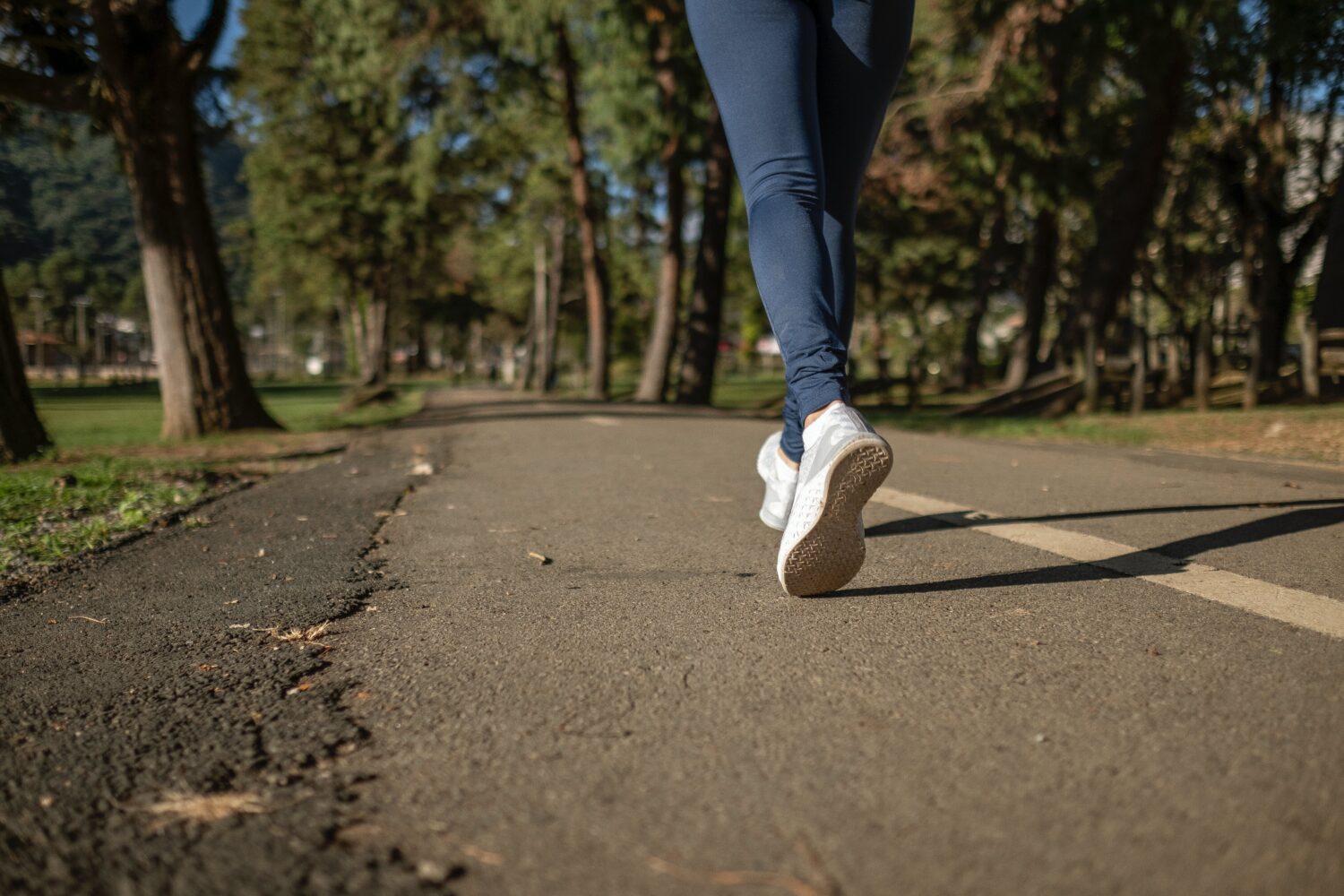 debloat, Woman wearing navy blue leggings and white and grey sneakers power walks on a paved outdoor path. 