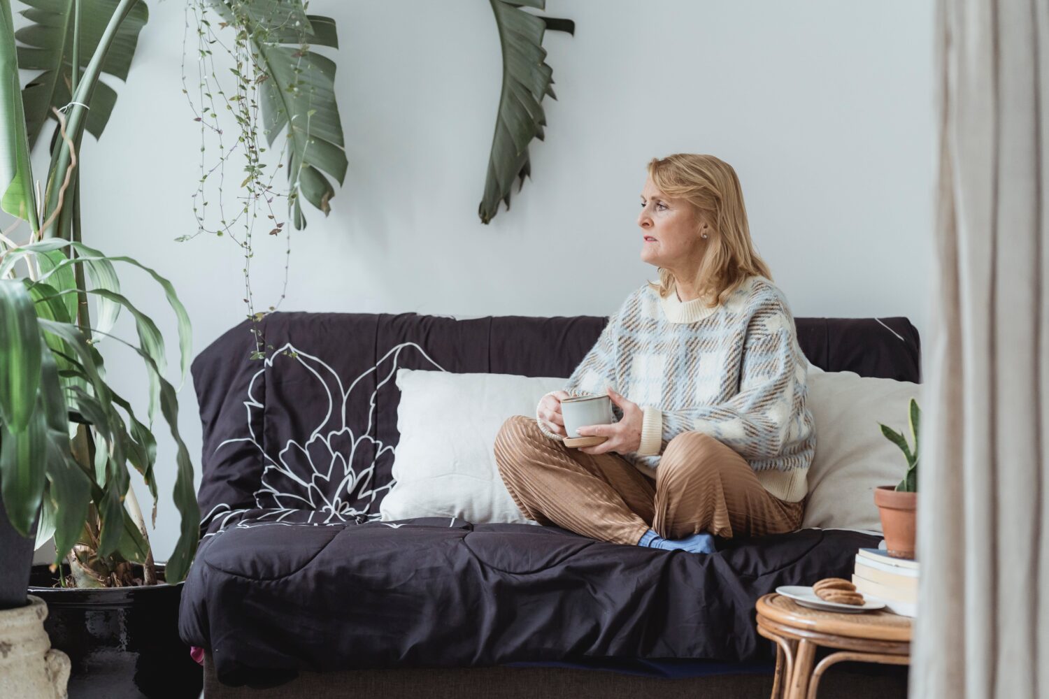 debloat, Blonde middle aged woman sits with her legs crossed and a cup of tea in her hands on a black couch with white pillows.