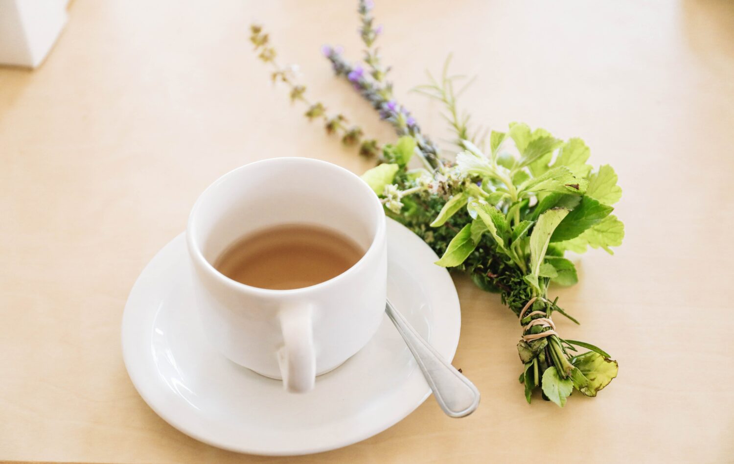 debloat, Half filled white tea cup with saucer and spoon sits on a wooden table next to a bundle of herbs.